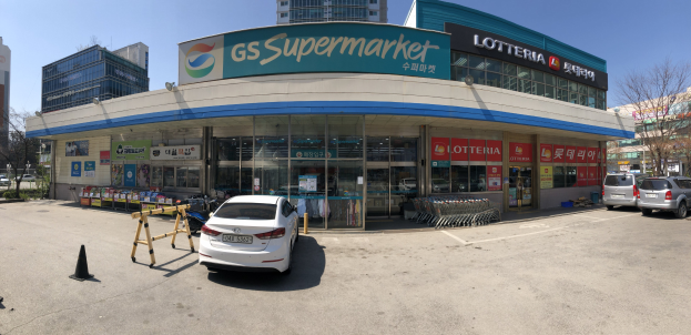 Ein Auto steht vor einem "GS Supermarkt"-Geschäft mit anderen Fahrzeugen, Verkehrskegeln und verstreuten Gegenständen in der Nähe, vor einem Gebäude, Bäumen und einem klaren blauen Himmel.