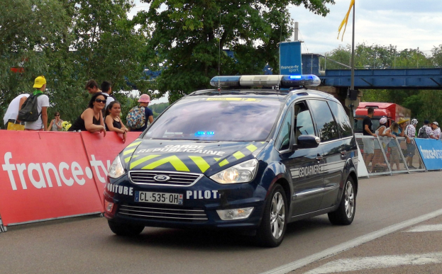Polizeifahrzeug fährt neben einer Menge mit Schildern, Geländern, Bäumen, einer Brücke, einer Flagge und einem bewölktem Himmel im Hintergrund.