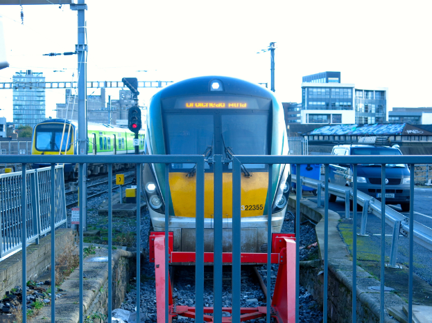 Zwei Züge auf den Schienen mit einem Zaun im Vordergrund, Pfosten mit einem mit einer Signallampe, ein Auto auf der Straße rechts, Gebäude im Hintergrund und Himmel darüber.