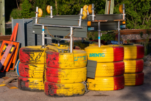 Ein Haufen gelber und roter Reifen auf einem Schotterfeld, umgeben von verschiedenen Gegenständen einschließlich Metallteilen und Brettern, mit Bäumen, Pflanzen und einer Hütte im Hintergrund.