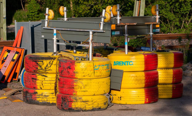Ein Haufen gelber und roter Reifen auf einem Schotterfeld, umgeben von verschiedenen Gegenständen einschließlich Metallteilen und Brettern, mit Bäumen, Pflanzen und einer Hütte im Hintergrund.