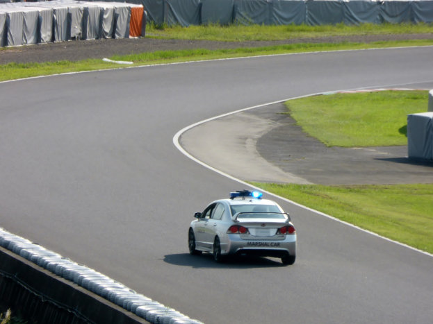 Polizeiauto fährt auf einer Rennstrecke mit Gras auf beiden Seiten und Zuschauern im Hintergrund.