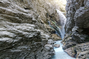 Kleiner Wasserfall, der über zerklüftete Felsen in einem steinigen Tal hinabstürzt, umgeben von grünen Hügeln, mit klarem Wasser und hellem Sonnenlicht, das die Szene erhellt.