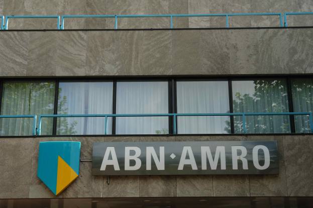 Ein Gebäude mit der Aufschrift "ABN AMRO" an der Seite, das eine Tafel mit der Aufschrift "Hauptsitz" zeigt, Geländer und Glasfenster mit Vorhängen, mit Text am unteren Rand, der eine Strafe von 1,5 Milliarden US-Dollar durch die Europäische Union erwähnt.