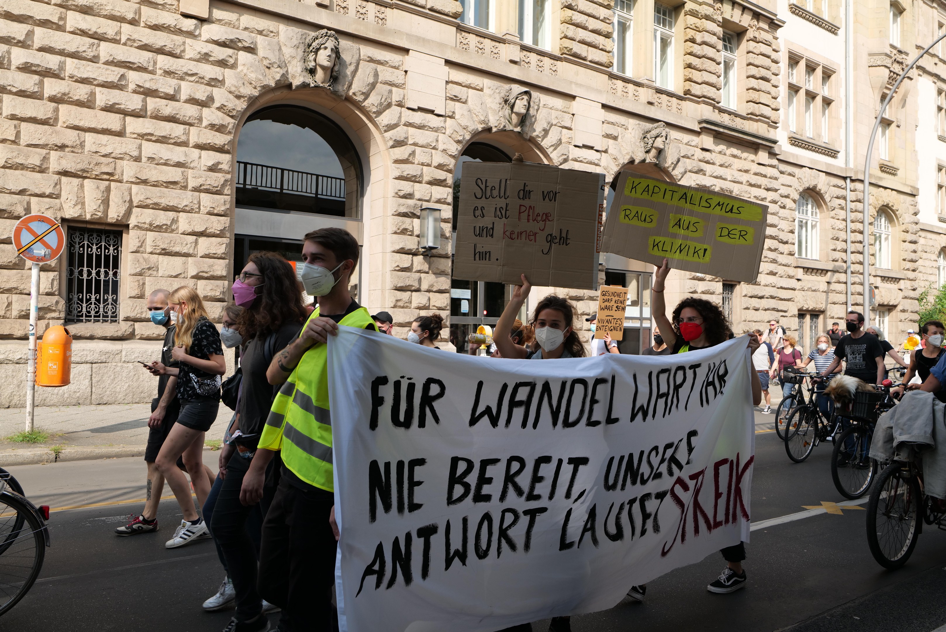 Eine Gruppe von Menschen, die auf einer Demonstration durch die Straße marschieren, einige halten Schilder und andere fahren Fahrräder, im Hintergrund ein Gebäude mit Fenstern, Bögen, Säulen und Skulpturen.