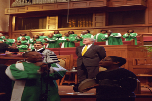 Eine Gruppe von Menschen steht vor einem Chor in einer Kirche, wobei eine Person in einem schwarzen Kleid ein Musikinstrument hält und eine andere in ein Mikrofon singt, während andere klatschen und im Hintergrund gegen braune Wände sitzen.