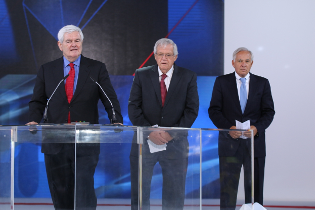 Ein älterer Mann in einem schwarzen Mantel und roter Krawatte steht an einem transparenten Rednerpult auf einer Bühne, mit einem blauen und weißen Banner im Hintergrund.