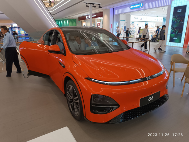 Oranger Elektroauto in einer Ausstellungshalle umgeben von Menschen, Stühlen, einem Wagen und Ladenschildern im Hintergrund.