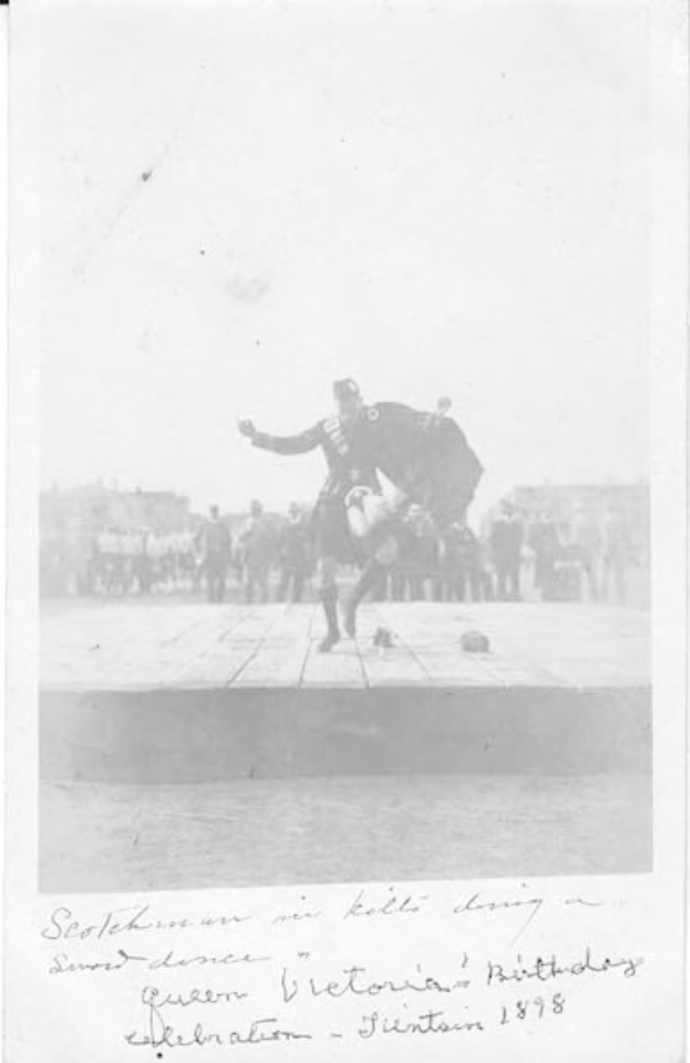 Schwarz-weißes Foto eines Mannes in Kilt, der auf einer Bühne tanzt, während der Geburtstagsfeier von Queen Victoria in Edinburgh 1898, mit einer Menge und Gebäuden im Hintergrund.