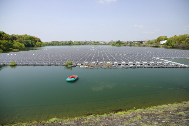 Ein kleines Boot schwimmt auf einem Gewässer, umgeben von grünem Gras und Bäumen, mit Gebäuden und einem klaren blauen Himmel im Hintergrund und Solarpanelen auf der Wasseroberfläche.