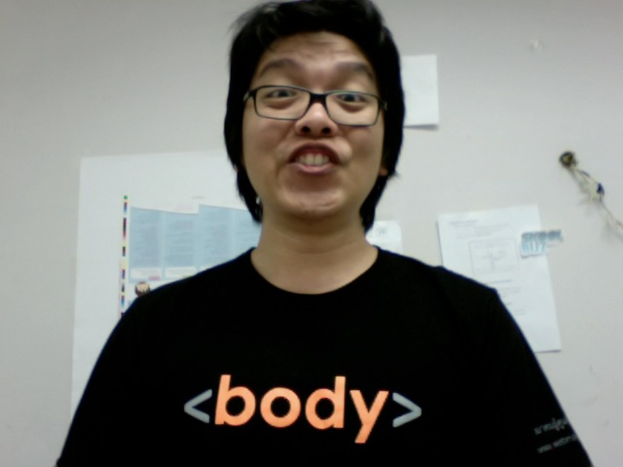 Eine Person in einem schwarzen T-Shirt und Brille lächelt in der Mitte des Bildes, mit Plakaten an einer weißen Wand im Hintergrund.