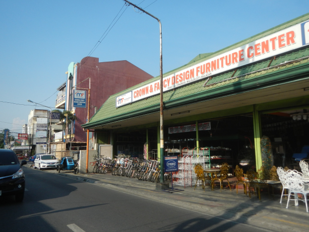Eine belebte Stadtstraße mit fahrenden Fahrzeugen, parkenden Fahrrädern, Gebäuden mit Schildern, Laternen und Stromleitungen, mit einem Möbelgeschäft im Vordergrund und einem sichtbaren Himmel im Hintergrund.