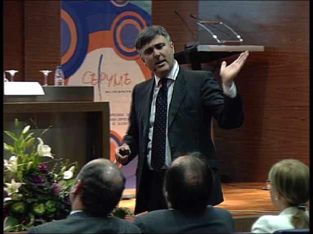 Zwei Männer und eine Frau sitzen auf Stühlen, während ein Mann in einem schwarzen Blazer an einem Podium mit Mikrofonen steht und in eines spricht, mit einem Blumentopf oder einem Strauß in der Nähe und einem Tisch mit einem Buch, einer Wasserflasche und anderen Gegenständen daneben, vor einer braunen Wand und einer bunten Tafel mit Text im Hintergrund, wahrscheinlich in einem Konferenzsaal.