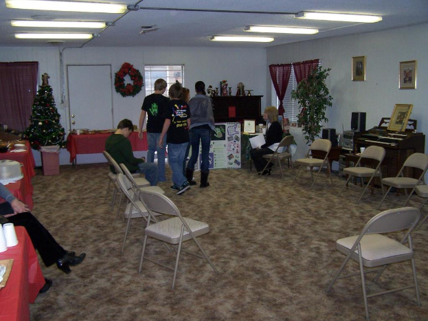 Ein Raum mit mehreren Stühlen, Tischen und Personen, einige stehen, einige sitzen. Auf den Tischen stehen Tassen und Gefäße. Im Hintergrund eine Wand mit einem Strauß, Vorhängen, einer Tür, Fotorahmen und Fenstern. Ein Weihnachtsbaum und ein Piano auf der rechten Seite. Röhrenleuchten an der Decke.