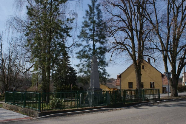 Ein Holocaust-Gedenkmonument steht in der Mitte einer Straße, umgeben von einem Metallzaun, umgeben von Grün, Häusern und einem bewölkten Himmel.
