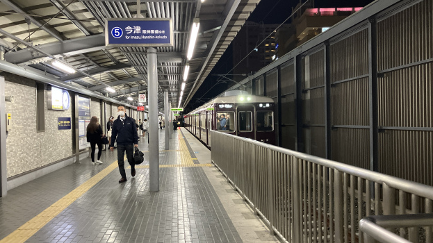 Ein Zug fährt auf einem Bahnsteig ein, mit Menschen in der Nähe, einem Geländer auf der rechten Seite, Gebäuden im Hintergrund, Bahnsteigbeleuchtung und einer Abfahrtsanzeige.