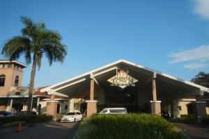 Der Eingang zu einem Resort mit Palmen vorne, umgeben von Gebäuden, Fahrzeugen auf der Straße, Verkehrskegeln, Büschen, Bäumen und einem bewölkten Himmel im Hintergrund.