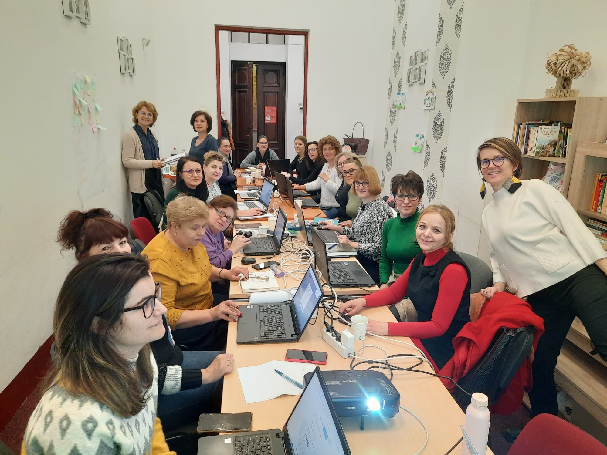 Eine Gruppe von Frauen mit Behinderungen nimmt an einem Workshop teil, die um einen Tisch mit Laptops, Papieren und anderen Gegenständen sitzen, mit Jacken auf Stühlen, einem Bücherregal und einer Vase und Papieren an der Wand im Hintergrund.