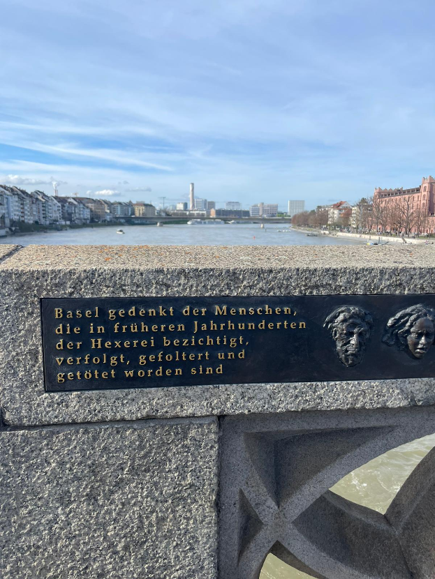 Eine Steinmauer mit einer Tafel, auf der "Basel Gedekt der Menschen" steht, neben einem Gewässer, mit Bäumen, Gebäuden und einem klaren Himmel im Hintergrund.