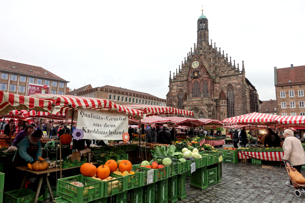 Ein belebter Markt in Nürnberg, Deutschland, mit verschiedenen Obst- und Gemüsesorten, einkaufenden Menschen, Zelten, Gebäuden mit Fenstern und einem Uhrenturm im Hintergrund unter einem sichtbaren Himmel.