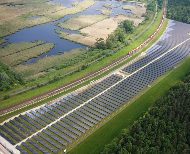Luftbild einer Solar-Farm mit Solar-Panelen, umgeben von Bäumen, Gras, Pflanzen und Wasser, mit einem Zug auf einer nahen Eisenbahnschiene.