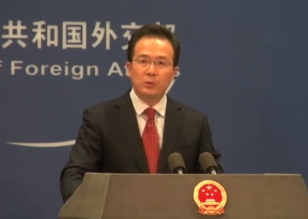 Ein Mann in Anzug und Krawatte spricht an einem Podium mit zwei Mikrofonen und einem Logo, vor einem Banner mit Text.