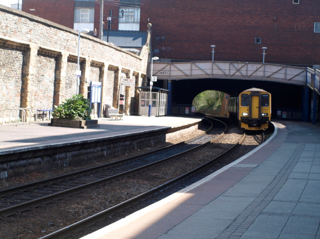 Ein Zug fährt auf Schienen mit Plattformen auf beiden Seiten, eine Hauswand links, Straßenlaternen, Pflanzen, Bänke, eine Hütte, eine Brücke über die Plattformen und Gebäude und Masten im Hintergrund.