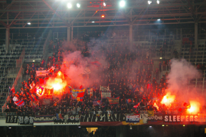 Eine große Menschenmenge in einem Stadion mit Feuerwerkskörpern und Rauch, die Fahnen und Schilder halten, unter einer Decke mit Deckenleuchten und umgeben von Metallrahmen und Pfosten.