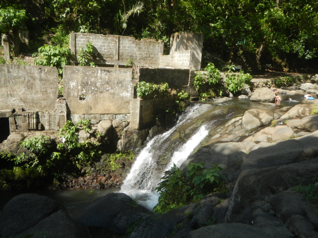 Ein kleiner Wasserfall ergießt sich über eine steile Felswand in einem üppigen grünen Wald, mit Menschen, die im Wasser schwimmen und einer Wand im Hintergrund.
