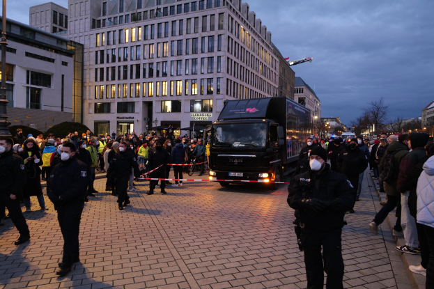 Eine Gruppe von Menschen steht vor einem Lastwagen auf einer Straße, umgeben von Gebäuden, Laternenpfählen, Bäumen und einem bewölkten Himmel, wobei einige Mützen und Masken tragen und ein Band mit einem Stab im Vordergrund zu sehen ist.