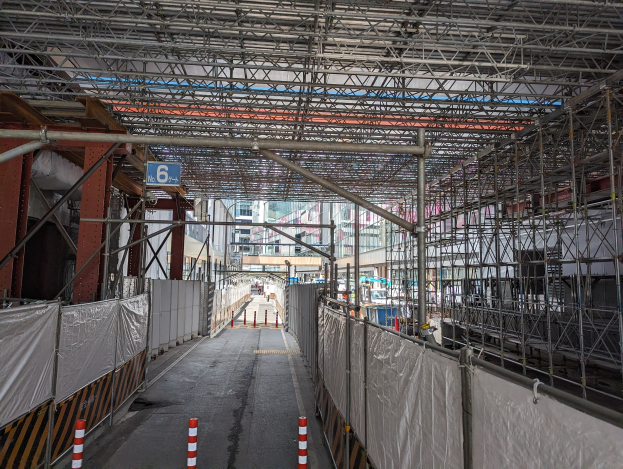 Eine Baustelle mit ausgedehnten Gerüten, darunter Stäbe, Metallrahmen und eine Tafel mit Text, neben einer Straße, einem Zaun und Gebäuden unter einem bewölktem Himmel.