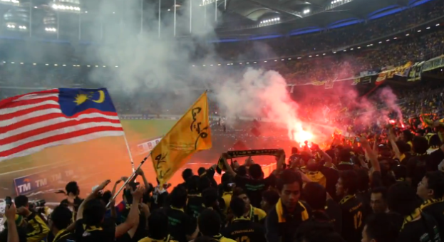 Eine große Menge Menschen in einem Stadion mit Fackeln und Rauch, einige halten Fahnen und eine Kamera, mit Schildern, die "Malaysia vs Malaysia Live Streaming" anzeigen.
