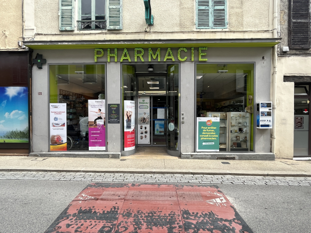 Außenansicht einer Apotheke mit Glaswänden und -türen, Plakate an den Wänden, an einer Straßenecke gelegen.