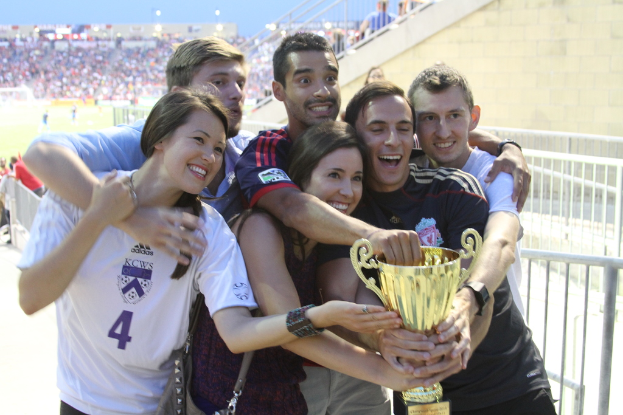 Sechs Menschen, zwei Frauen und vier Männer, halten einen Pokal und lächeln, posieren für ein Foto vor einem Eisenzaun, mit einer Wand und einem Zaun oben rechts, Menschen und Lichter im Hintergrund, möglicherweise in einem Stadion.