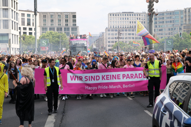 Große Gruppe von Menschen marschiert auf einer Straße mit einer "Happy Pride March Berlin"-Fahne, während ein geparktes Auto auf der rechten Seite und Gebäude, Bäume und ein klarer Himmel im Hintergrund zu sehen sind.