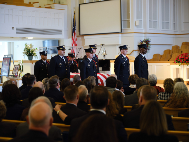 Gruppe uniformierter Personen vor sitzenden Trauernden bei einer Beerdigungszeremonie, mit Blumenarrangements, einer Fahne, einem Bildschirm und einer Wand im Hintergrund.