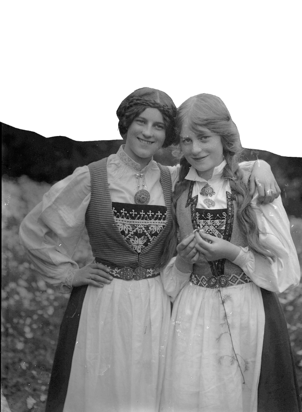 Zwei junge Frauen in traditioneller bayrischer Tracht, mit weißen und schwarzen Kleidern mit aufwändiger Stickerei und Perlen, posieren vor der Kamera vor einem Hintergrund aus üppigen grünen Pflanzen und einem hellen weißen Himmel.