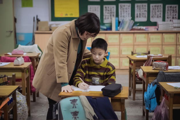 Eine Frau und ein Junge sitzen an einem Tisch in einem Klassenzimmer, wobei der Junge in ein Buch schreibt.