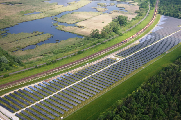 Luftaufnahme eines Solarparks mit Panelen in einer grünen Wiese, umgeben von Bäumen und Wasser, mit einem Zug auf einer nahen Bahntrasse.