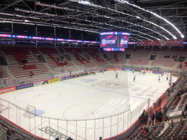 Eishockeyspiel in einer großen Arena zwischen den St. Louis Blues und Detroit Red Wings, mit Zuschauern auf Sitzen und Stehplätzen in der Nähe der Eisfläche, umgeben von einem Zaun, einem Bildschirm, Metallrahmen und Deckenleuchten.