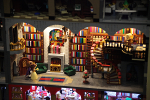 Ein Lego-Modell einer Bibliothek mit Bücherregalen, einer Treppe, einem Kronleuchter, einem Kamin, einer Sitzgruppe, einem Tisch, Stühlen, einem Teppich und verstreuten Spielzeugen.