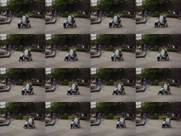 Ein Kind geht auf einer Straße und zieht einen Wagen, mit zwei Personen, die links Fahrräder halten, Bäumen im Hintergrund und einem Gebäude links im Bild.