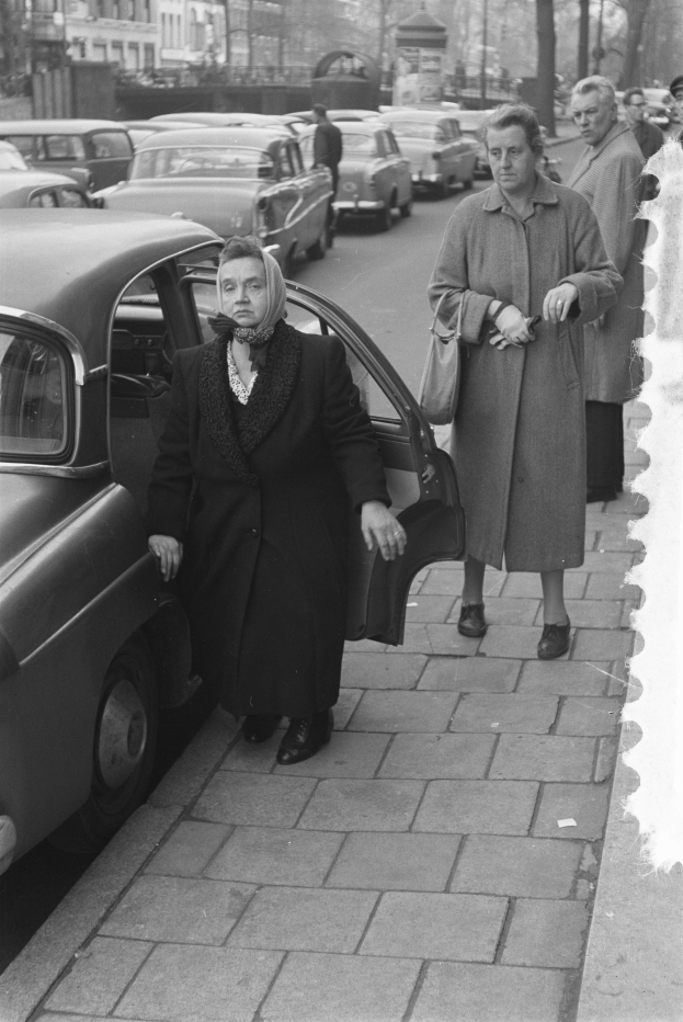 Eine Schwarz-Weiß-Szene einer Frau, die aus einem Auto auf einer Stadtstraße aussteigt, mit anderen Fahrzeugen, Passanten, Bäumen, Polen, Gebäuden und einem klaren Himmel im Hintergrund.