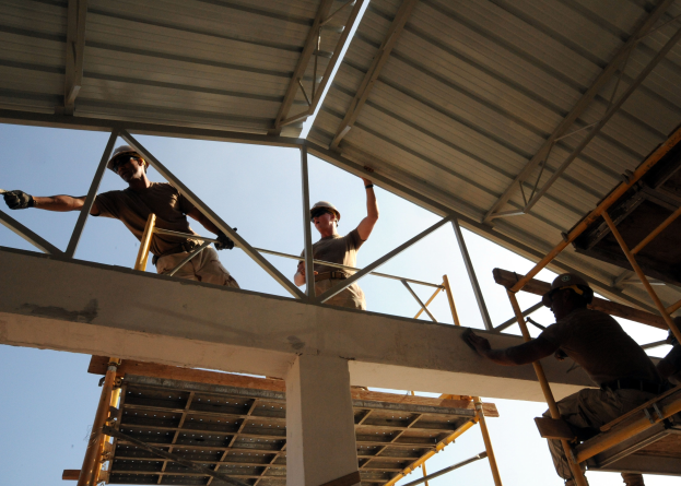 Drei Männer mit Helmen und Handschuhen arbeiten auf dem Dach eines Gebäudes unter Bau, stehen auf Leitern und halten Stangen, mit einer Säule im Vordergrund und dem Himmel im Hintergrund.