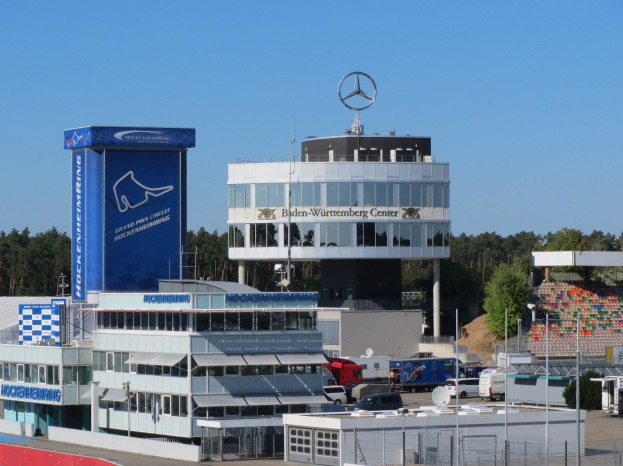 Ein Blick auf den Mercedes-AMG Petronas F1 Team Hauptsitz in Berlin, Deutschland, mit Gebäuden, Fahrzeugen auf der Straße, einem Zaun, Pfählen, Schildern mit Text, Bäumen und einem Logo auf dem Gebäude, mit dem Himmel im Hintergrund.