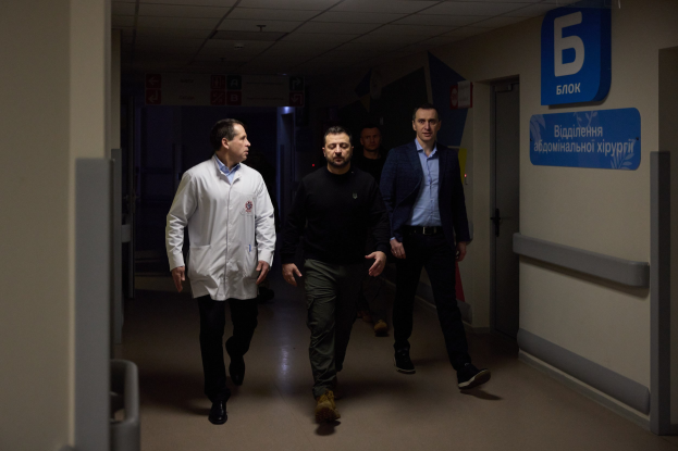 Eine Gruppe von Männern geht einen Krankenhausflur entlang mit Deckenleuchten, einer Tür an der rechten Wand und Texttafeln an der linken Seite, die besorgt aussehen.