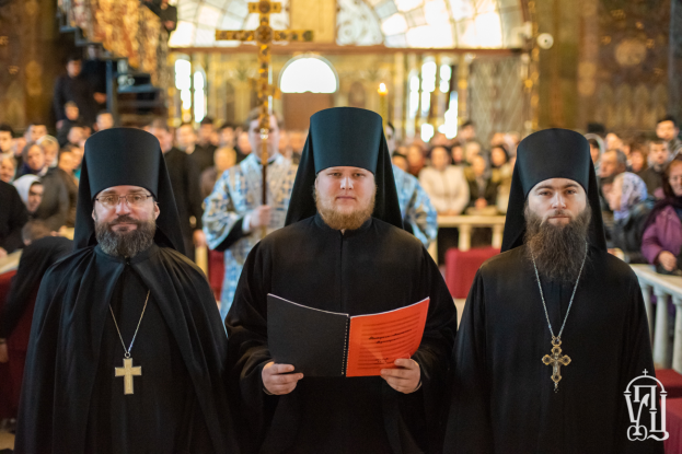 Drei Priester in schwarzen Gewändern und Hüten stehen vor einer Menschenmenge in einer Kirche, einer hält ein Buch, mit einem unscharfen Hintergrund und einem Wasserzeichen in der rechten unteren Ecke.