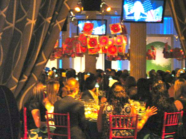 Eine Gruppe von Menschen, die an Tischen mit Essen, Fernsehern, Lampen, hängenden Papieren und Vorhängen in einem Raum sitzen.