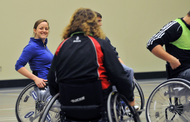 Eine Gruppe von Menschen in Rollstühlen auf dem Boden sitzend, mit einer Frau in einer blauen Jacke und einer anderen Person in einem grünen Hemd, vor einer Wand im Hintergrund.