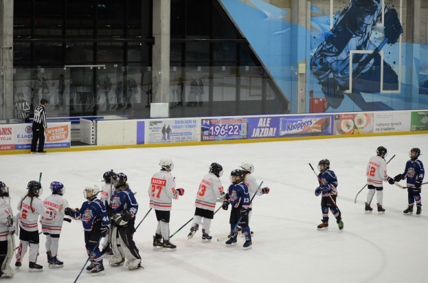Gruppe junger Menschen, die Eis-hoc-key auf einer Indoor-Eisfläche spielen, mit Helmen und Stöcken, während eine Person links steht und Banner, Säulen und Glaswände im Hintergrund zu sehen sind.